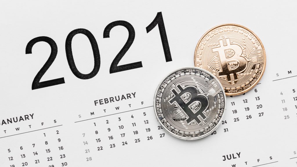 calendário do ano de 2021 com duas moedas colocadas por cima dela, uma prata e outra bronze, ambas estão com um cifrão em formato de B no centro indicando serem uma analogia a moedas digitais